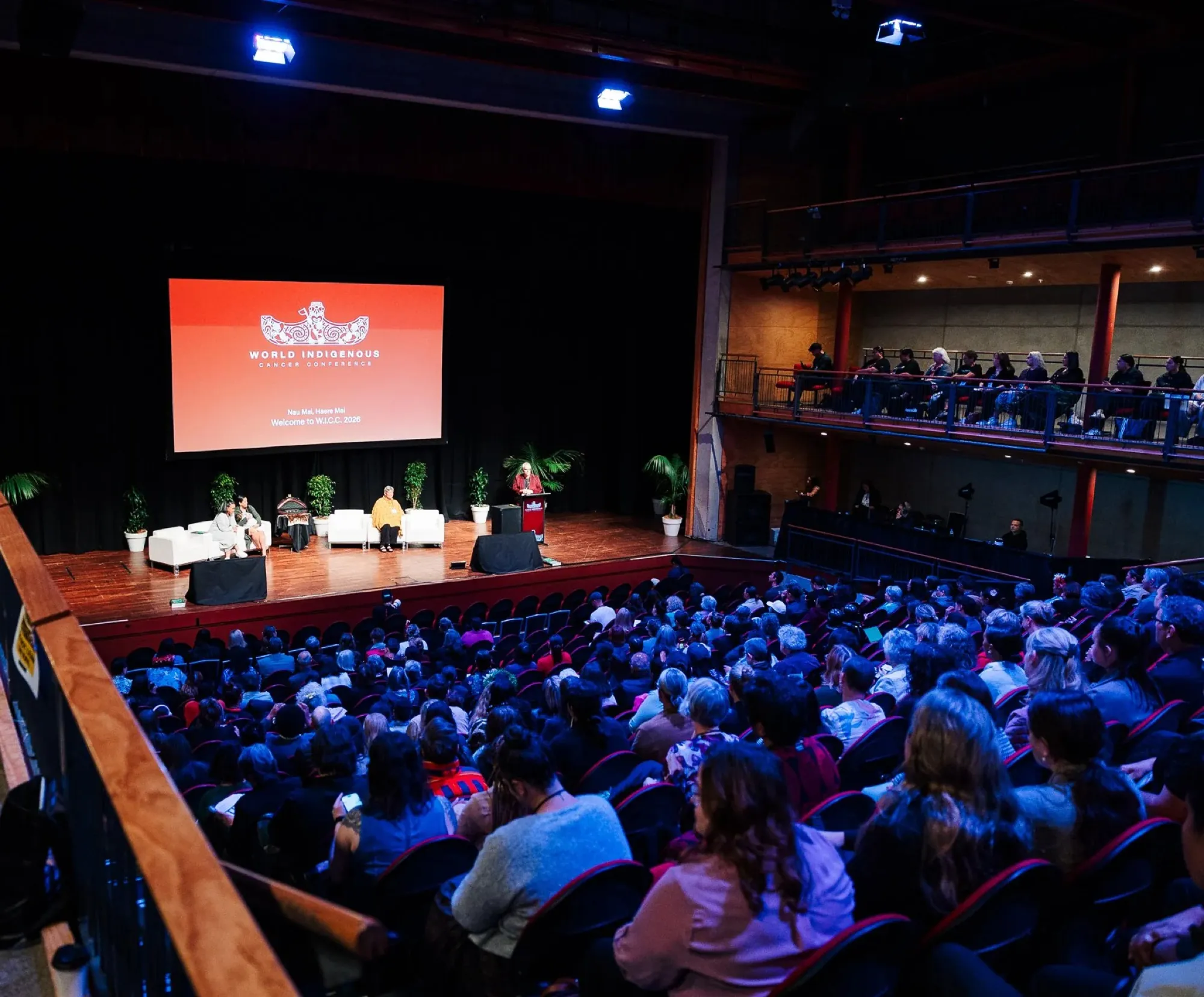 A full theatre facing the stage and the World Indigenous Cancer Conference screen during a plenary session, Tāmaki Makaurau, April 2026